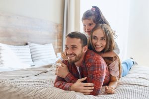 Positive optimistic man lying on bed in comfortable hotel room, keeping hands crossed over, looking away, wife and child happily resting on his back, holiday time concept, shot from below